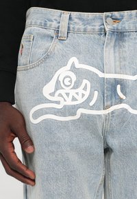 Light blue denim jeans with a relaxed fit, featuring a large white graphic design of a cartoon cat on the front. Subtle stitching details.