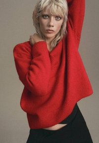 Pull en laine rouge avec un col en V, coupe ample et poignets côtelés, associé à une ceinture noire, sur un fond neutre.