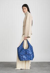 Μπλε τσάντα ώμου με σχέδιο, σε χαλαρή σχήμα, κατασκευασμένη από ανθεκτικό υλικό. Διαθέτει ευρύχωρο σχεδιασμό με μεγάλη άνοιξη και στιβαρές λαβές.