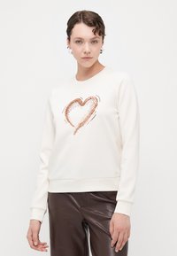 Kremgenser med et hjerte-design i rosegull-glitter, med rund hals og ribbestrikkede ermer, parret med brune lærbukser.