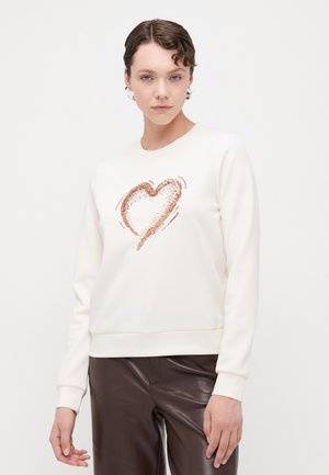 Kremgenser med et hjerte-design i rosegull-glitter, med rund hals og ribbestrikkede ermer, parret med brune lærbukser.