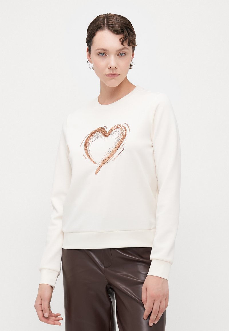 Kremgenser med et hjerte-design i rosegull-glitter, med rund hals og ribbestrikkede ermer, parret med brune lærbukser.