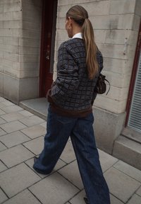 Hnedý pletený kardigán s geometrickými vzormi, prehodený cez tmavú košeľu, v kombinácii s voľnými modrými džínsami. Doplnok: malá hnedá taška.