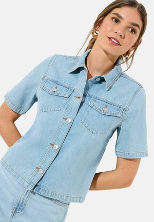 Noor naine, kes kannab helerohelist lühikeste varrukatega teksasärki ees taskutega ja sobivaid teksapükse, poseerib valge tausta ees.