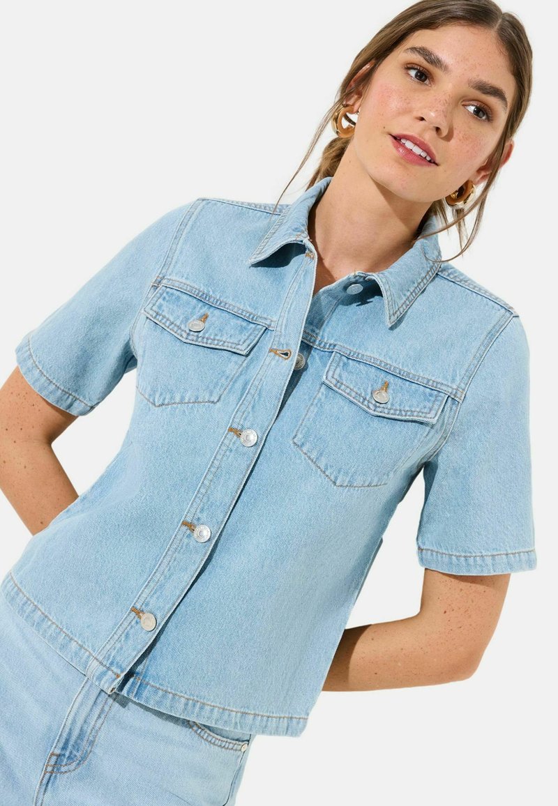 Noor naine, kes kannab helerohelist lühikeste varrukatega teksasärki ees taskutega ja sobivaid teksapükse, poseerib valge tausta ees.