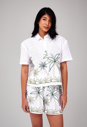 Vrouw die een witte set draagt met een kort shirt met korte mouwen en korte broek, bedrukt met een groen en beige palmboom- en graspatroon, staand tegen een effen achtergrond.