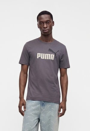 Jeune homme à la peau foncée portant un t-shirt gris Puma et un jean bleu clair à motif Puma, debout devant un fond uni.