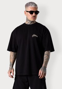 Schwarz oversized T-Shirt aus Baumwolle, mit einem beige gestickten Logo auf der Brust. Glatte Textur mit lockerer Passform und kurzen Ärmeln.