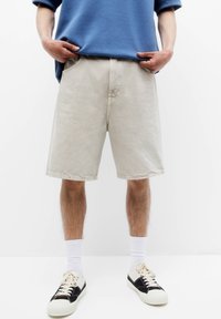 Hombre que lleva pantalones cortos beiges hasta la rodilla, calcetines blancos, zapatillas negras y blancas, y una camisa azul con las manos en los bolsillos.