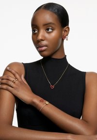 Conjunto de joias apresentando um colar de corrente dourada com um pendente em forma de coração vermelho, brincos combinando e vários anéis com designs minimalistas, numa modelo.