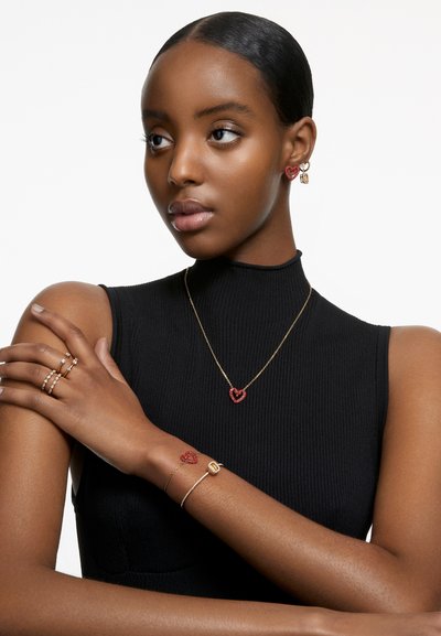 Ensemble de bijoux comprenant un collier en chaîne dorée avec un pendentif en forme de cœur rouge, des boucles d'oreilles assorties et divers bagues à designs minimalistes, porté par un modèle.