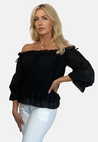 Czarny top z odkrytymi ramionami, z falbaniastymi rękawami, elastyczną talią i warstwowym dołem, uszyty z lekkiego materiału. Połączony z białymi dżinsami.