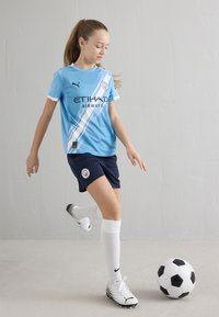 Šviesiai mėlynas futbolo marškinėlis su baltais akcentais, tamsiai mėlynos kelnės ir balti kojinės. Žaidėjas veiksme su juodai balta futbolo kamuoliu.