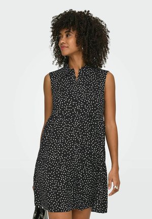 Kvinne med krøllete hår iført en svart ermeløs kjole med hvite prikker, holder en svart håndveske, ser til siden.