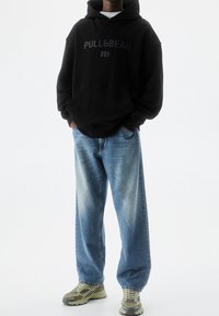 Personne portant un sweat à capuche noir avec le texte « PULL&BEAR », un jean bleu à coupe large et des chaussures de sport beige-vert, debout les mains dans les poches.