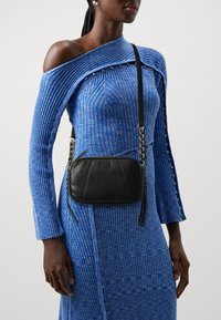 Borsa a spalla in pelle nera con zip, tracolla intrecciata e dettaglio di nappina, indossata accanto a un vestito blu a coste con un design unico.
