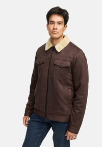Braune Kunstlederjacke mit einem cremefarbenen Schaffellkragen, Reißverschluss vorne, zwei Brusttaschen und weicher Textur. Getragen mit blauen Jeans.
