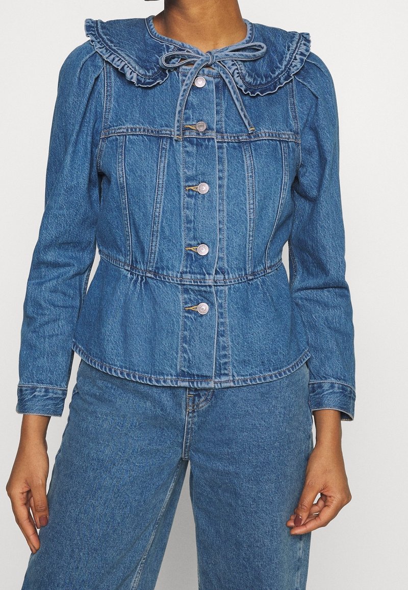 Denim jacket with a ruffled collar, button front, fitted waist, and long sleeves, featuring a light blue wash and subtle texture details.