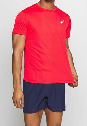 Mann trägt ein leuchtend rotes, kurzärmeliges Sportshirt und dunkelblaue Shorts, hält mit einer Hand den Saum des Shirts vor einem schlichten hellen Hintergrund fest.