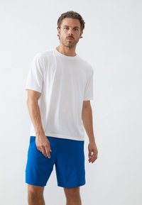 Λευκό T-shirt με κοντό μανίκι και στρογγυλή λαιμόκοψη, σε συνδυασμό με μπλε σορτς. Το ύφασμα φαίνεται απαλό, με χαλαρή εφαρμογή.