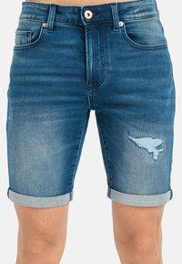 Pantalones cortos de mezclilla en azul medio con un parche desgastado y puños enrollados. Presenta cinco bolsillos y una cremallera con un cierre de botón metálico.