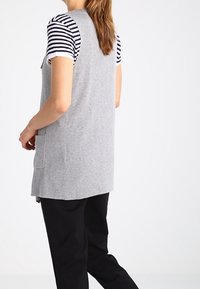 Šedá pletená vesta s bočními kapsami, nošená přes pruhované tričko s krátkým rukávem. Design má volný střih a střední délku.