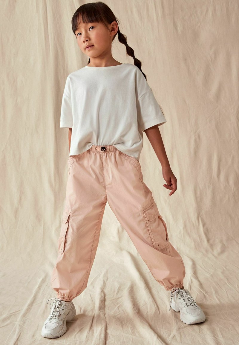 Lichtroze cargobroek met elastische boorden en zakken, gecombineerd met een witte t-shirt met korte mouwen. Model draagt witte sneakers en staat tegen een beige achtergrond.