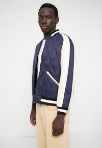 Jaqueta bomber acolchoada em azul-marinho e creme, com punhos canelados, fechos de correr, bolsos laterais e um design de risca contrastante nas mangas.