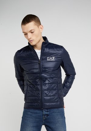 Giacca imbottita blu navy con design trapuntato, chiusura con zip, colletto rialzato e logo EA7 ricamato sul petto. Indossata sopra una camicia a righe.