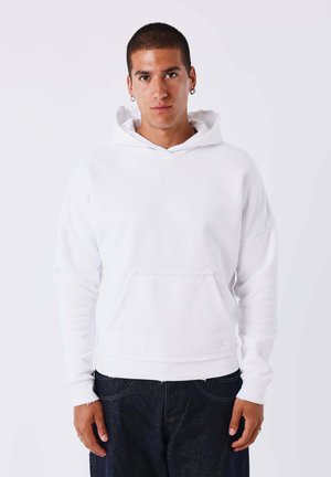 Jonge man met kort haar, gekleed in een witte hoodie en donkere spijkerbroek, staand tegen een effen lichte achtergrond.