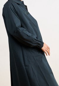 Granatowy płaszcz z kołnierzykiem, długimi rękawami i elastycznymi mankietami. Z przodu znajduje się kieszeń, a materiał jest gładki i lekki.