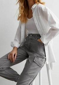 Frau in einem weißen ärmellosen Top und einem oversized weißen Hemd mit Knöpfen, trägt hochgeschnittene graue Cargo-Hosen und sitzt auf einem weißen Hocker.