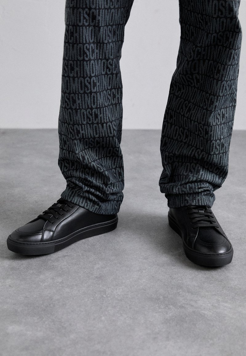 Black leather sneakers with a textured finish, featuring a round toe and flat rubber sole, paired with patterned dark fabric pants.