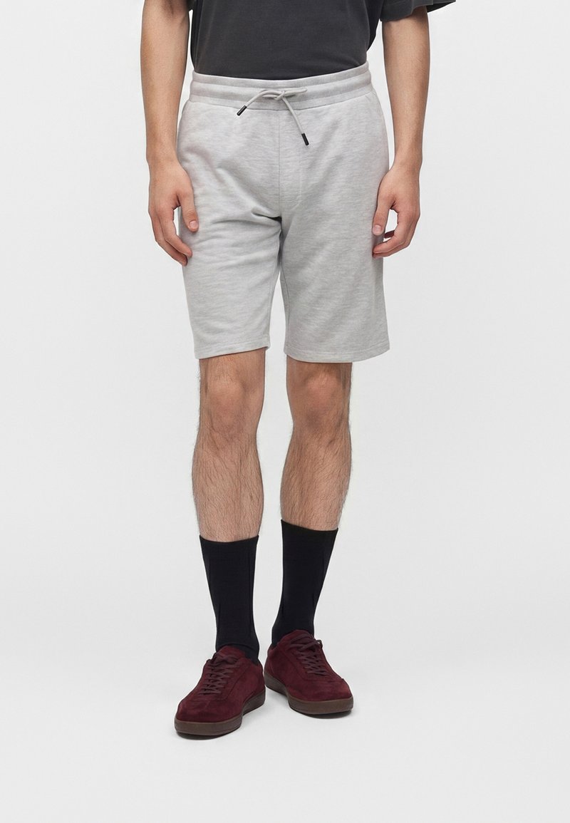 Personne portant un short gris clair avec cordon, des chaussettes noires mi-mollet et des baskets rouge foncé, debout devant un fond uni.