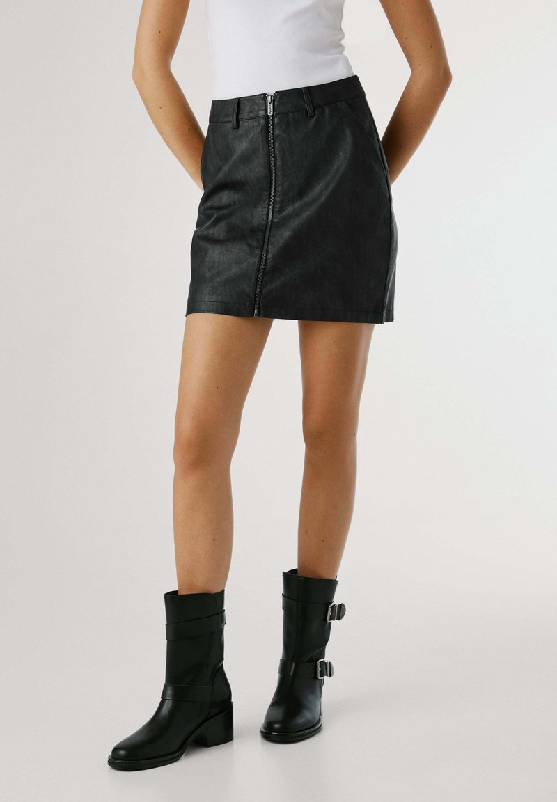 Gonna mini nera in finta pelle con zip frontale, vita piatta e design senza cuciture. Modella con stivaletti neri con tacco.