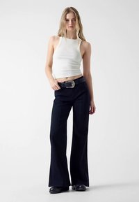 Débardeur blanc ajusté associé à un pantalon noir taille haute et évasé. Le pantalon a une texture lisse et une coupe large. Détail boucle de ceinture argentée.
