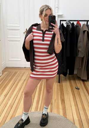 Mujer con un vestido de rayas rojas y blancas con cuello marrón, sosteniendo una chaqueta marrón sobre los hombros, tomando una selfie frente al espejo en el interior.