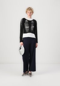 Schwarzer, strukturierter Strickpullover mit Ausschnitten, über einem weißen Oberteil getragen, kombiniert mit navyblauen, weit geschnittenen Hosen und silbernen Accessoires.