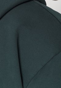 Tmavě zelená mikina s kapucí, charakterizovaná žebrovanou strukturou, měkkou tkaninou a bez viditelných log nebo ozdob. Detaily švů zdůrazňují design.