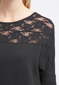Top nero con scollatura in pizzo floreale, tessuto morbido e vestibilità rilassata. La blusa ha maniche lunghe e un semplice colletto tondo.