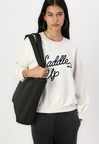 Felpa bianca con testo ricamato scuro, abbinata a una borsa a tracolla nera. Tessuto texturizzato, vestibilità comoda e polsini a costine. Pantaloni grigio scuro.