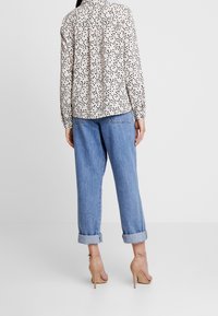 Femme portant un chemisier beige à manches longues avec des pois noirs, un jean bleu clair retroussé et des sandales à talons hauts couleur nude, debout devant un fond blanc.