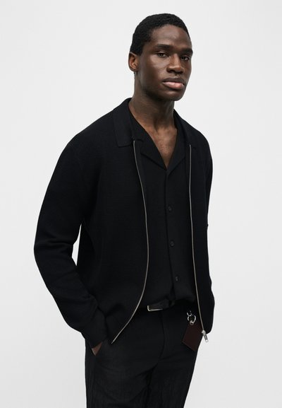 Νεαρός άνδρας που φοράει μαύρο υφασμάτινο σακάκι με φερμουάρ πάνω από μαύρο πουκάμισο με κουμπιά, με τα χέρια στις τσέπες, στέκεται μπροστά σε ένα απλό φωτεινό φόντο.