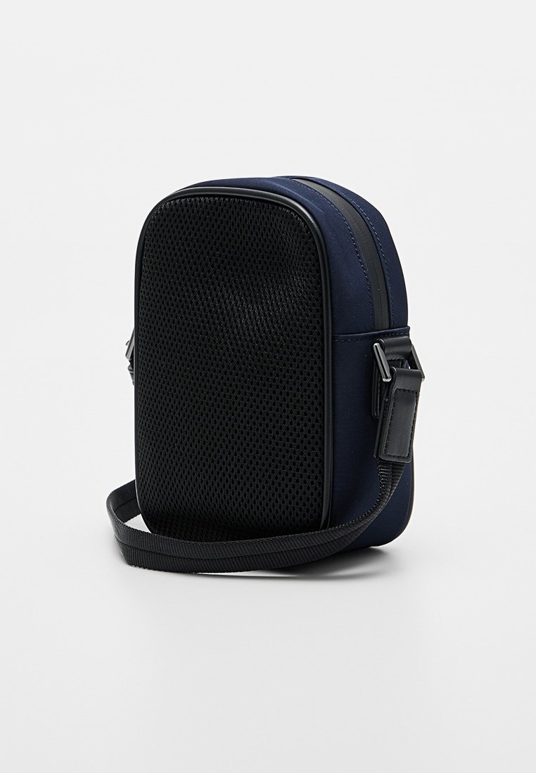 Bolso de hombro azul marino con un frente de malla negra texturizada, forma rectangular, correa ajustable y detalles de herrajes negros.