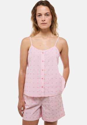 Femme aux cheveux châtain clair ondulés portant un haut rose à pois sans manches boutonné et un short assorti, debout avec une main dans sa poche.