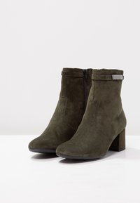 Botines de ante en verde oscuro con punta redonda, tacón de bloque y cremallera lateral. Presentan un puño doblado y un sutil acento metálico.