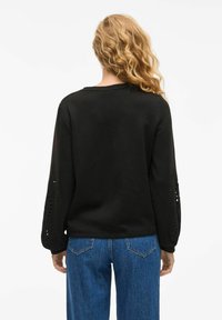 Felpa nera con maniche lunghe, caratterizzata da disegni floreali a taglio, scollatura rotonda e vestibilità rilassata, abbinata a jeans blu.