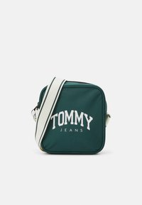Zelená čtvercová kabelka přes rameno vyrobená z nylonu, s bílým logem "TOMMY JEANS" na přední straně a nastavitelným proužkovaným popruhem.