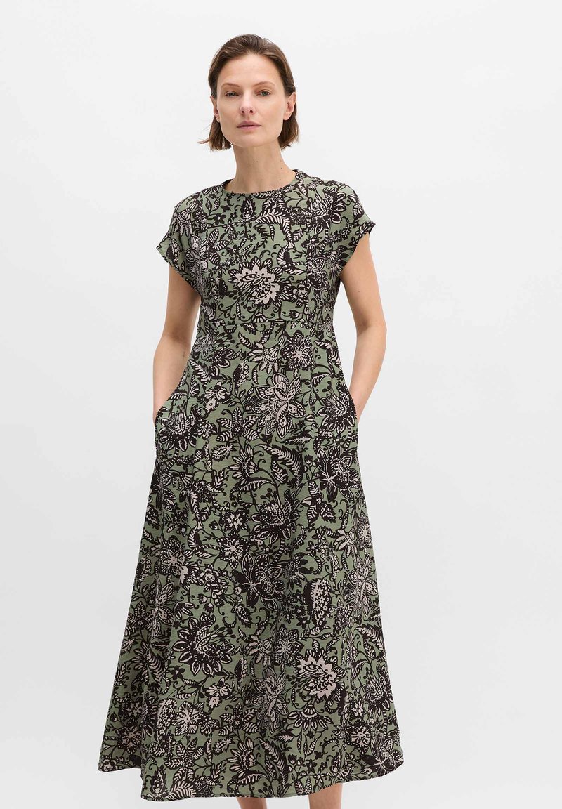 Frau steht mit den Händen in den Taschen und trägt ein grünes, geblümtes Midikleid mit kurzen Ärmeln und rundem Ausschnitt.