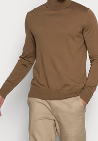 Brun polotröja i slät stickad tyg, med lång ärmar och ribbade muddar, parat med ljusbeiga byxor.